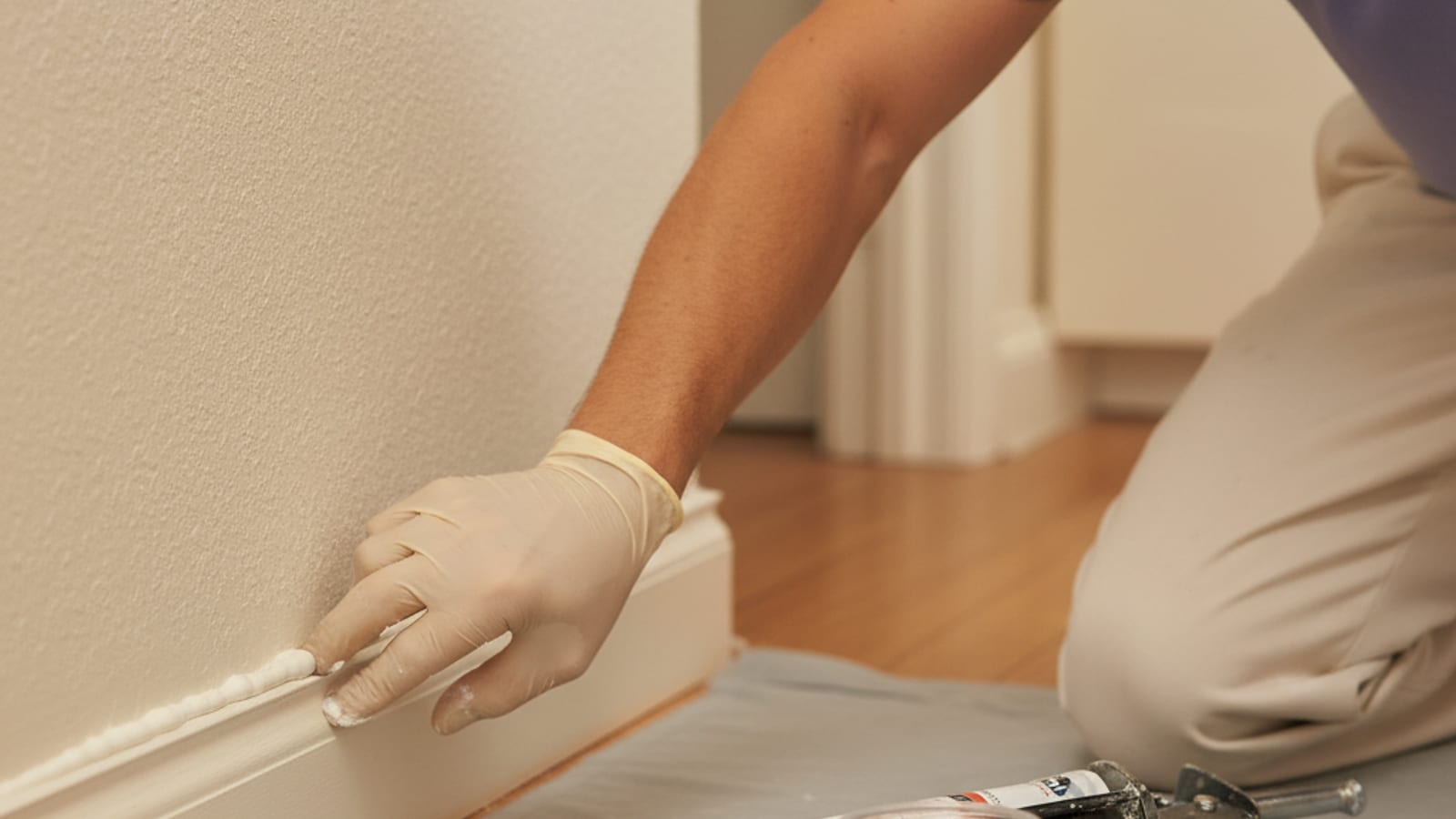 Hands smoothing a fresh bead of acrylic caulk along a baseboard-to-wall seam with a wet finger