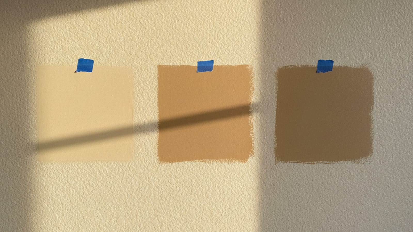 Three paint sample squares painted directly on a wall labeled with small blue tape tags